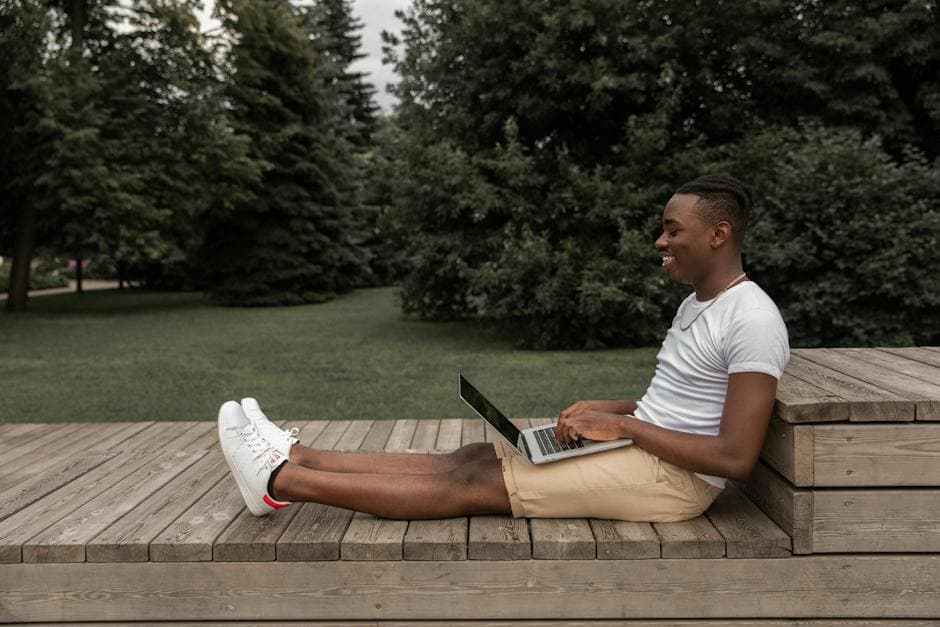 Side view of African American male in casual outfit sitting on wooden surface and browsing netbook while looking at screen