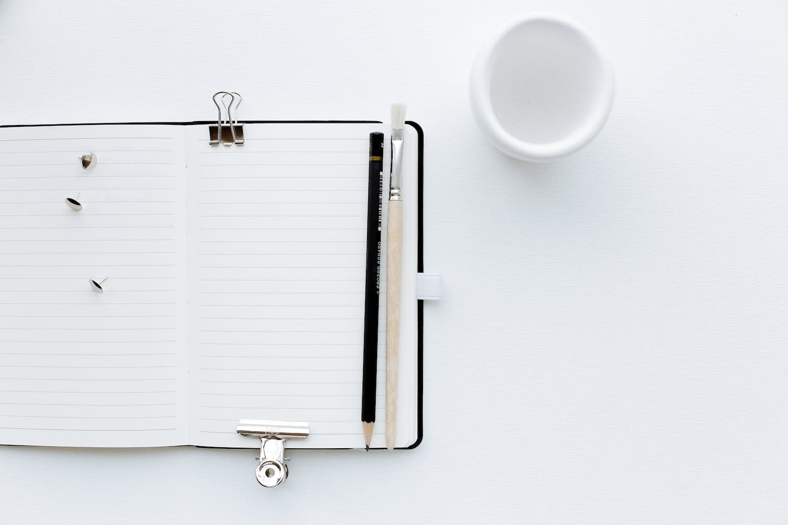Top view composition of opened diary with blank pages placed on white desk with pencil and paintbrush pins